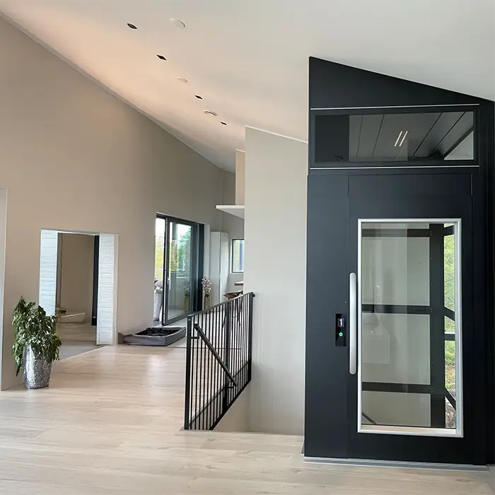 This platform lift installation shows the landing door with a full clear glass panel on the top landing. The lift structure is steel with solid steel panels, and the landing door has a brushed steel D handle.